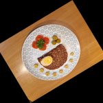 Auf einem gemusterten Teller befinden sich eine Scheibe Vollkornbrot, Tomaten, Oliven und Maiskölbchen