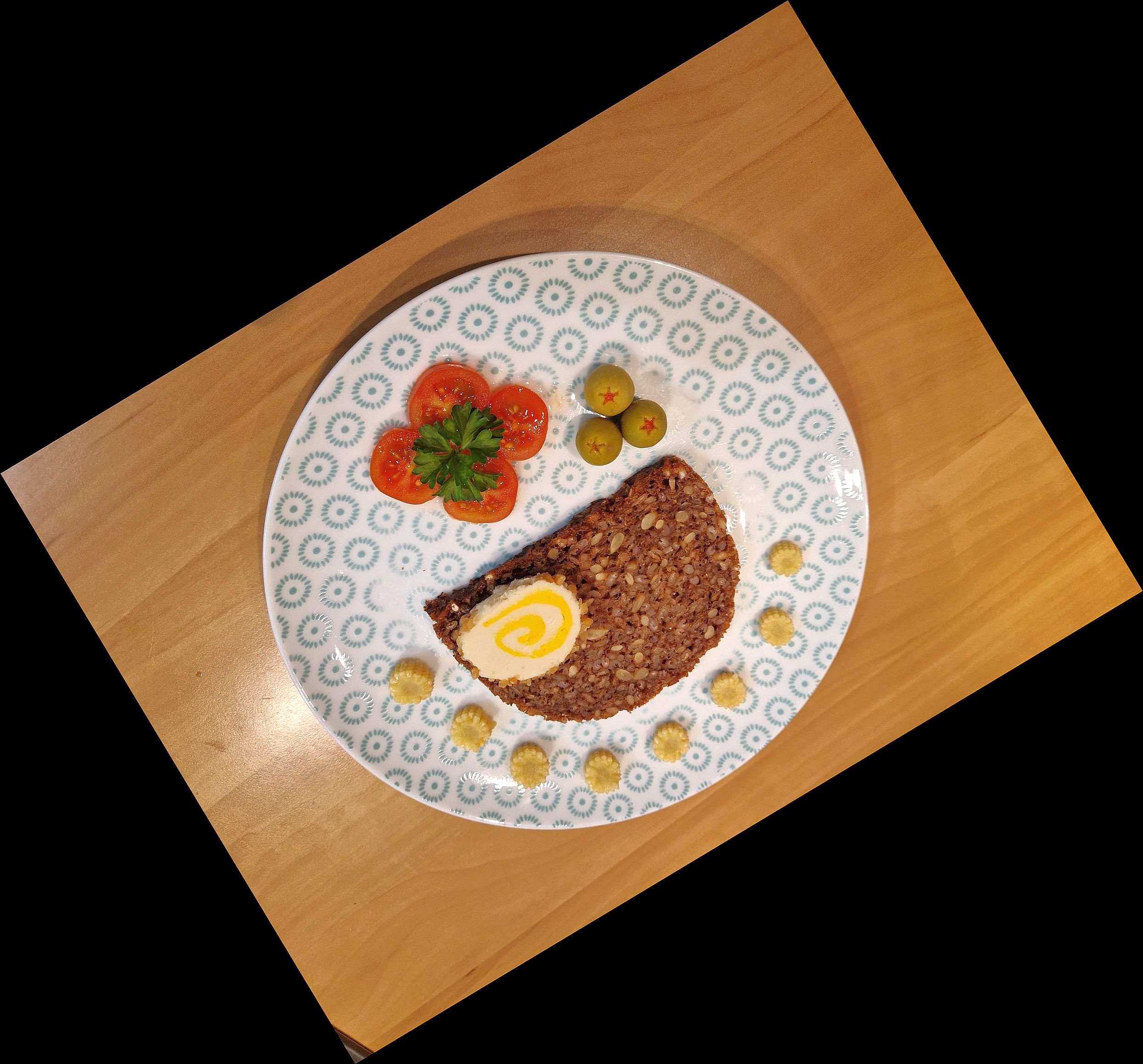 Auf einem gemusterten Teller befinden sich eine Scheibe Vollkornbrot, Tomaten, Oliven und Maiskölbchen