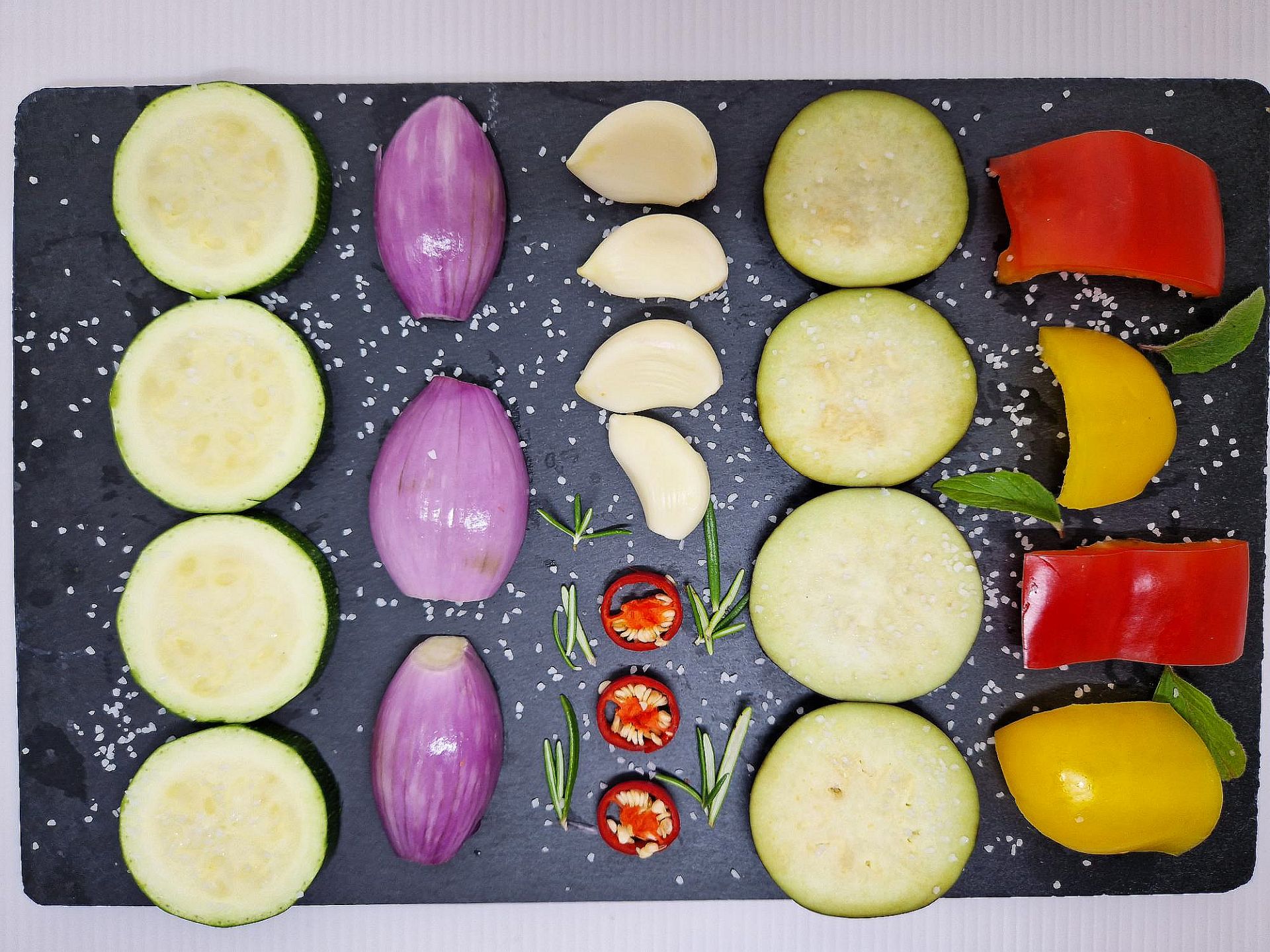 Zucchinischeiben, Schalotten, Knob lauchzehen, Chiliringe, Auberginenscheiben, Paprikastücke und abgezupfte Kräuter auf schwarzem Schieferbrett